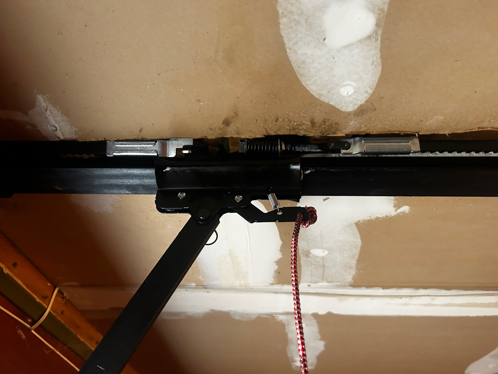 A black metal garage door opener rail and carriage assembly mounted to a ceiling with a red emergency release rope hanging down from the mechanism.