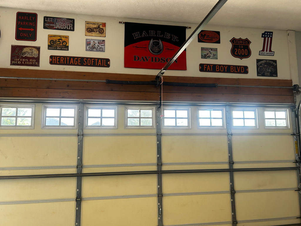 A garage wall with ten vintage-style Harley-Davidson signs above a closed garage door with rectangular windows.