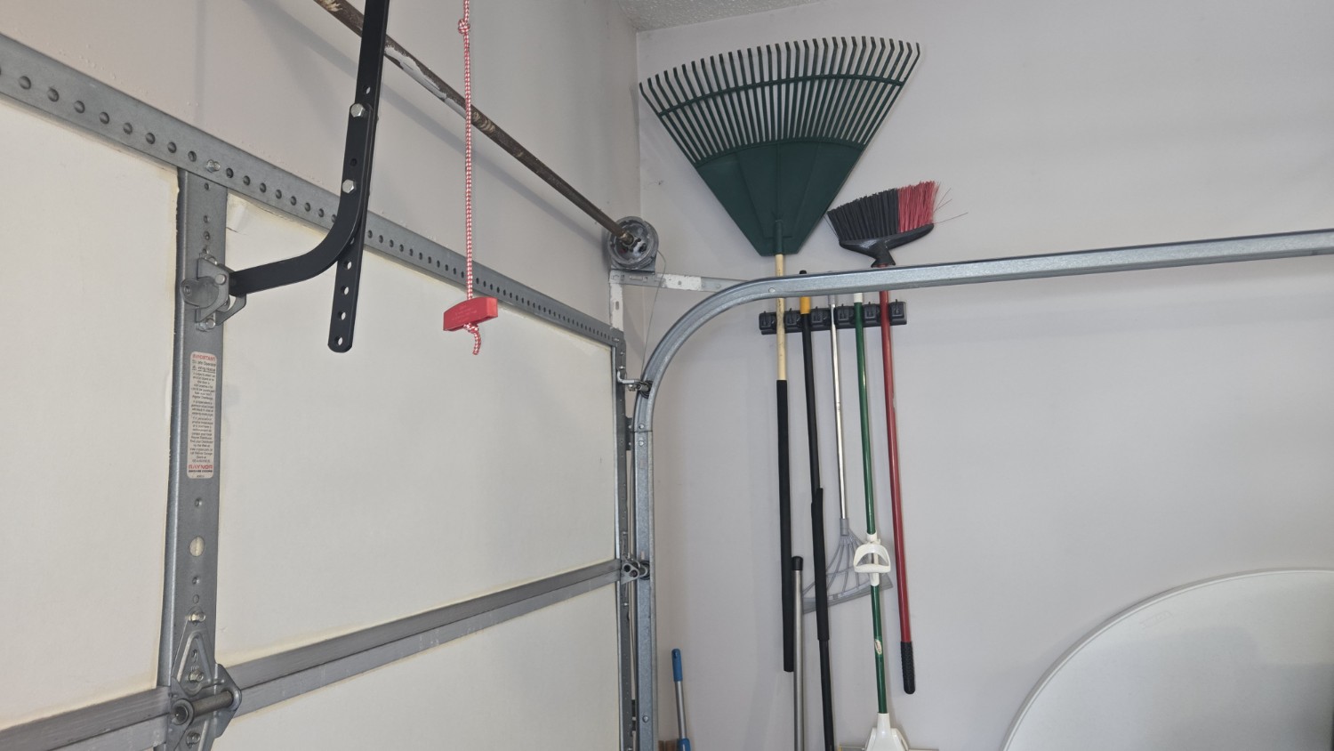 A tidy garage corner shows a fan rake, small brush, and long-handled garden tools hanging beside metal garage-door tracks, creating an orderly feel.